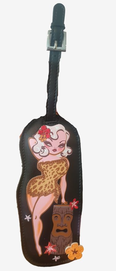 A luggage tag featuring an illustrated character resembling a cartoon leopard-print wearing woman, named Cavegirl Carlita, attached to a strap.