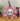 A pink luggage tag with a skull and crossbones design, featuring a blank address label on the back, displayed on a wooden stand.