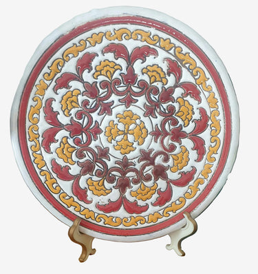 A vintage handpainted ceramic plate with earthy tones, featuring a floral pattern in golden yellow, maroon, brown, and terracotta on a white background, displayed on a stand.