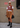 A woman stands on a city sidewalk wearing a white t-shirt, a rust-orange patterned knit vest, a brown pleated skirt, black boots, and sunglasses, holding a black handbag.