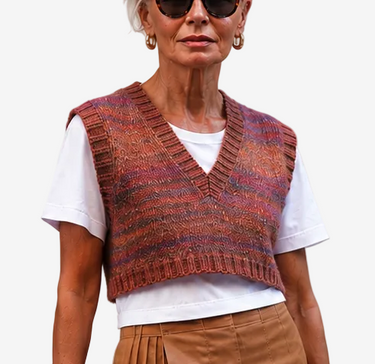 An older woman stands against a light gray studio background wearing a rust-brown knit vest over a white T-shirt, a brown wrap-style skirt, black ankle boots, and holding a small black handbag.