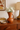 Terracotta vase with white flowers on a wooden surface, with a lamp and framed art in the background.