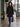 A woman walking on a city sidewalk wearing a black mid-length coat with a dark fuzzy hood, blue jeans, and black ankle boots, while carrying two paper shopping bags.