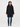 A woman modeling a black hooded coat with a fuzzy dark hood, paired with blue jeans and black ankle boots, against a plain light gray studio background.
