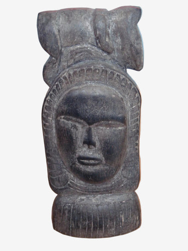 A dark carved wooden figurine of a stylized face with a headdress, standing on a light wood surface against a blue background.