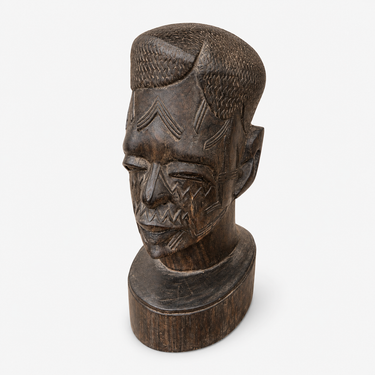 A dark carved wooden sculpture of a stylized human head with geometric facial patterns and a short textured hairstyle, mounted on a matching oval base against a plain white background.