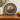 A round decorative ceramic plate with a hand-painted bird design, displayed on a wooden table in a styled home setting next to a vase with greenery and a bowl of pears.