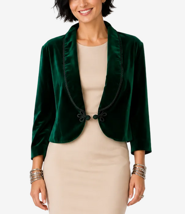 A woman modeling a dark green velvet cropped jacket over a beige dress, standing against a light gray studio background.