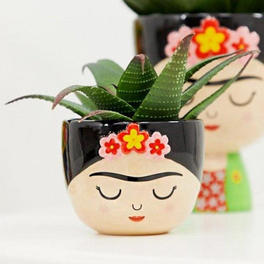 A small planter in the shape of a character resembling Frida Kahlo, with a black, white, and multicolor design, containing a green aloe plant.