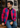 A man wearing a vibrant pink and blue checkered bomber jacket, a black t-shirt, and blue jeans, sitting on a wooden bench in front of a stone building.