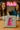 Two women sitting at a table with a pink dalek wallet in front of them, and a neon sign in the background.