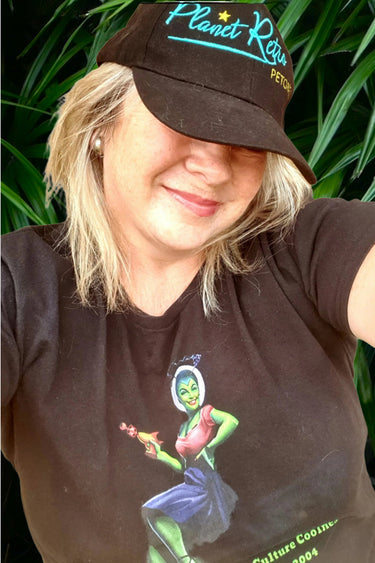 Person wearing a brown cap and t-shirt with a graphic design, standing against a green leafy background.