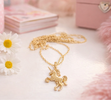 Gold unicorn necklace on a white surface with flowers and pink plush toys in the background