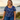 Woman wearing a blue floral top and jeans sitting on a wooden bench by the beach.