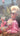 Lady in a colourful outfit sitting in a garden with Mount Taranaki, a UFO, and pink smoke in the background.  Planet Retro NZ.