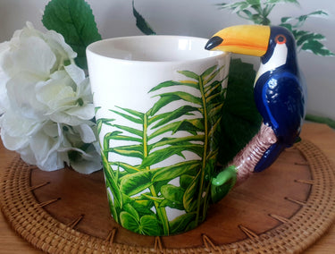 A white ceramic coffee mug with a green tropical plant pattern and a 3D toucan bird handle on a wooden surface, with a white flower in the background.