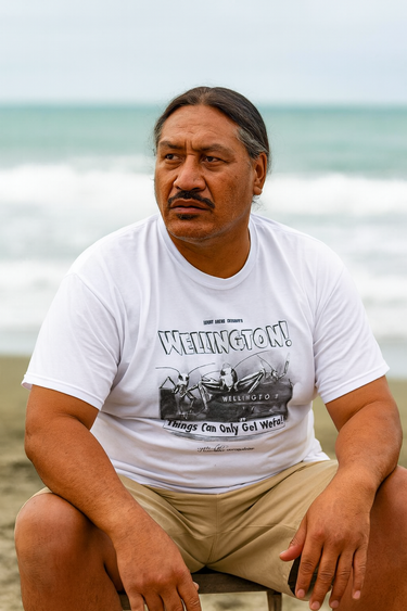Man sitting on a beach wearing a white t-shirt with text and graphics.