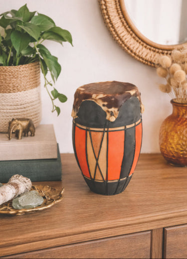 Decorative drum with orange and black design on a wooden surface, surrounded by plants and decor items.