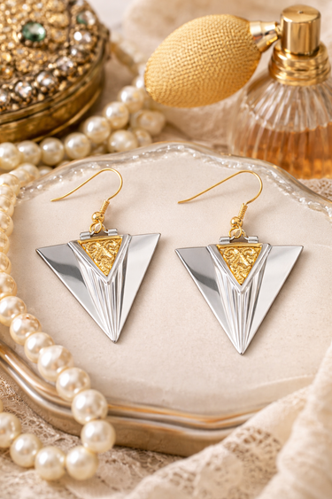 Pair of silver and gold triangle earrings on a decorative surface with pearls and a perfume bottle.