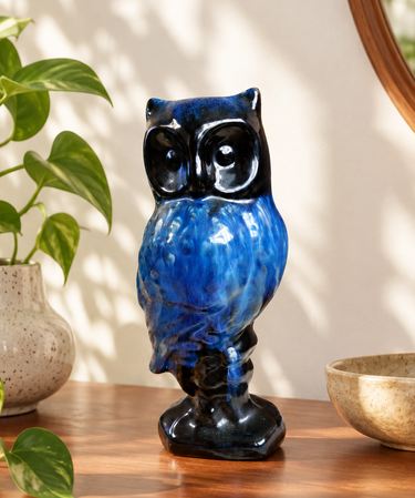A glossy Blue Mountain Pottery ceramic owl figurine with a vibrant blue and black glaze, standing on a small pedestal base displayed on a wooden console table next to a potted green plant and a bowl.