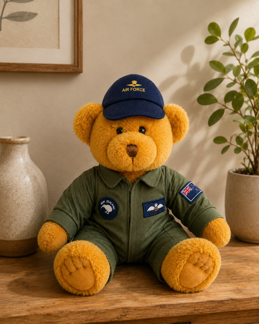 A plush teddy bear wearing an olive green flight suit and a navy blue cap with 'AIR FORCE' embroidered on it, sitting on a wooden surface in a styled indoor setting with a vase and potted plant.