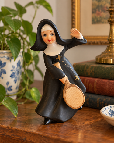 A vintage-style figurine of a female nun in a black habit holding a wooden tennis racket, displayed on a wooden table next to a potted plant and books.