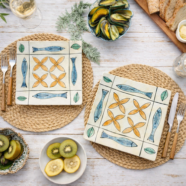 “Bird’s-eye view of a pair of Diana Poor New Zealand hand-decorated square ceramic plates with fish and floral motifs, styled on a rustic table setting.