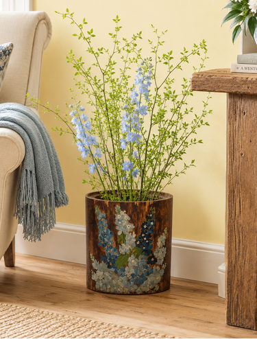 A rustic brown ceramic planter with a blue and white floral pattern, filled with green stems and light blue flowers, styled in a cozy living room next to a beige armchair and wooden side table.