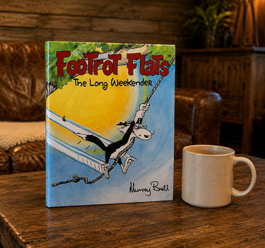 The book 'Footrof Flats: The Long Weekender' by Murray Roth stands open on a rustic wooden table next to a white ceramic mug, set against a cozy living room background with leather armchairs and wood paneling.