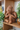 Inalco Terracotta sculpture of a seated figure on a wooden shelf with plants in the background