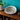 A large natural abalone shell with iridescent pearly tones sits on a wooden shelf in a styled interior setting, with a potted plant, books, and ceramic vessels visible against a teal wall.
