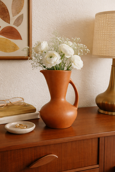 Terracotta vase with white flowers on a wooden surface, with a lamp and framed art in the background.