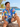 Male model wearing Lowes blue pineapple and hibiscus Hawaiian shirt with denim shorts, seated on driftwood beside a tropical beach