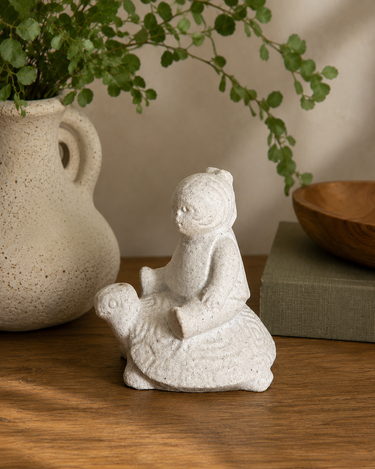 Decorative stone figure of a child on a turtle with a plant and wooden bowl in the background.