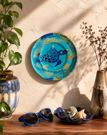A round ceramic wall plate featuring a blue sea turtle design with gold marbled accents, displayed on a textured beige wall above a wooden surface styled with sunglasses, seashells, and ceramic vases.