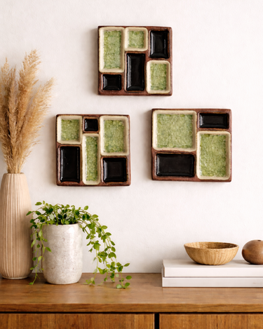 Three square ceramic wall art pieces on a white wall with a wooden cabinet and decorative items below.