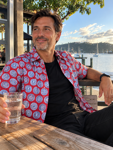 Man wearing red Wheels Planet Retro Original cotton shirt open over black T-shirt with black jeans, seated at waterfront cafe holding a glass of water