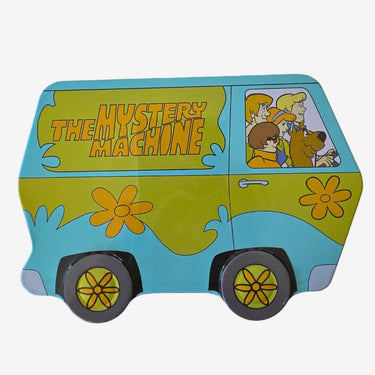 A toy or model of 'The Mystery Machine' van, featuring a light blue body with yellow flower decals, a yellow side panel with 'THE MYSTERY MACHINE' text, and cartoon characters in the window, displayed on a white textured fabric background.