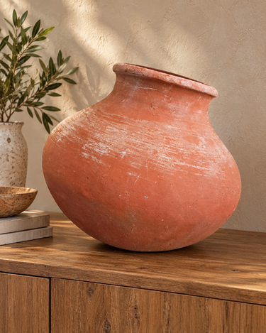 Rustic rounded terracotta vessel styled on a timber sideboard against a warm plaster wall with soft greenery
