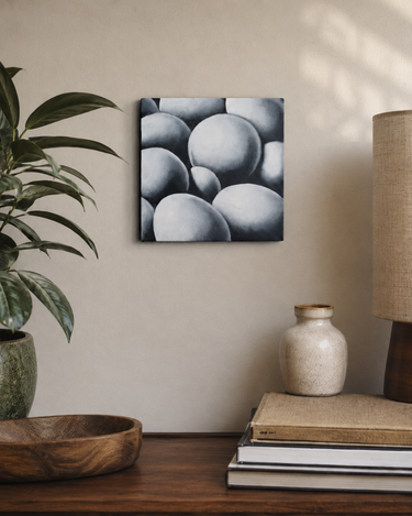 A square canvas wall art print featuring a monochrome painting of overlapping spheres, styled on a beige wall above a wooden table with a plant, books, a ceramic vase, and a lamp.