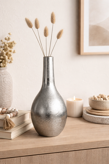 Silver vase with dried plants on a wooden surface with books and decor items.