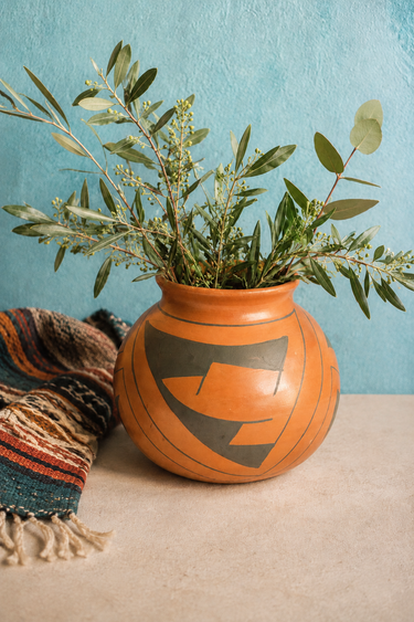 An orange clay vase with a black geometric pattern, filled with green olive branches, set on a beige surface against a textured light blue wall with a striped throw blanket on the left.