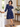Woman wearing Trollied Dolly navy blue swallows retro dress with bow neckline and navy heels, posing beside a staircase in a bright interior