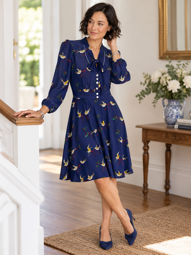 Woman wearing Trollied Dolly navy blue swallows retro dress with bow neckline and navy heels, posing beside a staircase in a bright interior