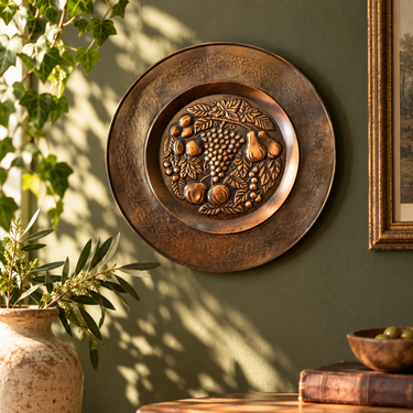 Vintage embossed copper fruit wall plate displayed on an olive green wall with dappled sunlight and soft foliage shadows