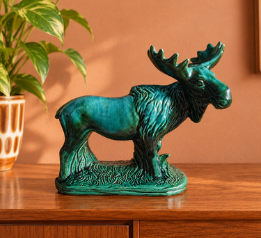 Vintage green glazed ceramic moose figurine displayed on a mid-century wooden sideboard against a soft terracotta wall with a leafy houseplant