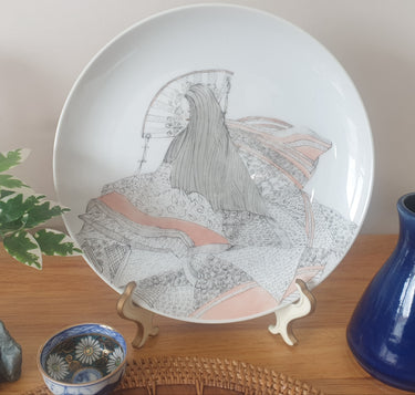 A hand-painted porcelain plate featuring a detailed painting of a long-haired Japanese maiden with peach highlights, signed by Joyce Humphrey, displayed on a wooden surface with a blue vase and a small decorative bowl in the background.