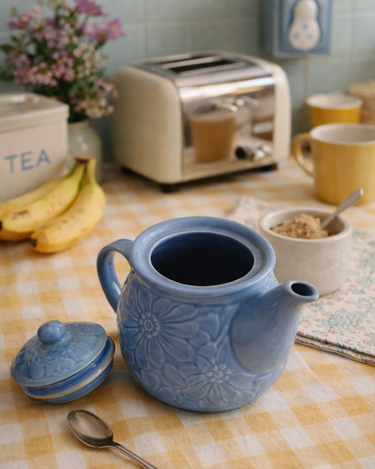 Blue ceramic teapot on a checkered tablecloth with a toaster and other kitchen items in the background.